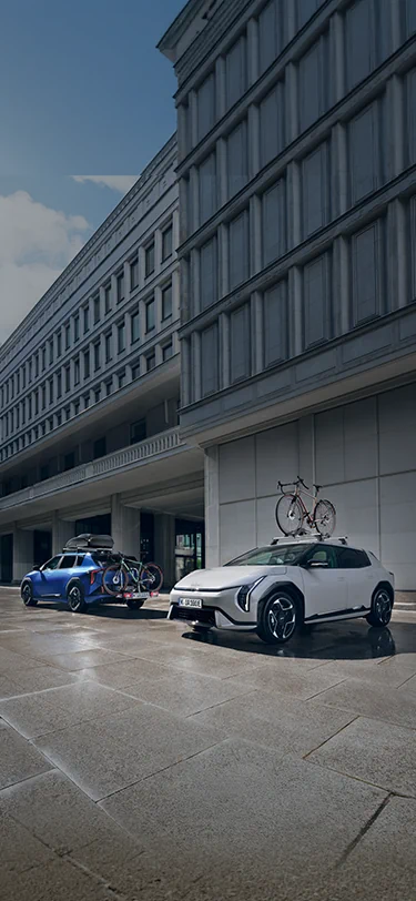 Zwei Kia EVs mit Kia Dachbox und Kia Fahrradträger Plus parken vor einem Gebäude.